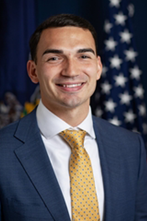 Man sitting in front of an American flag smiles at camera. He is wearing a navy suit, whit shirt, and yellow tie.